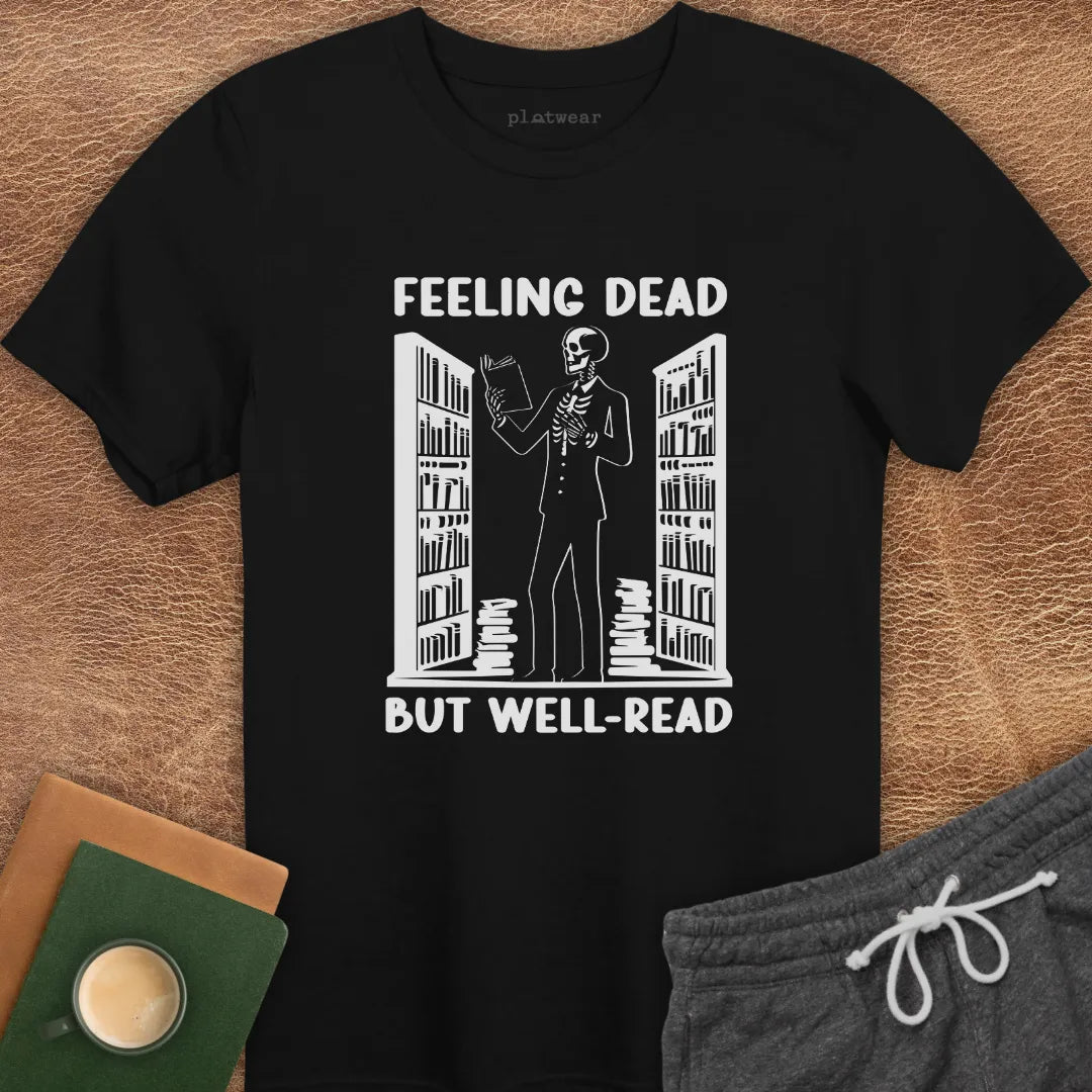 White crew neck t-shirt with the text 'FEELING DEAD BUT WELL-READ' printed on the front, displayed flat with a pair of gray shorts, a book, and a cup.