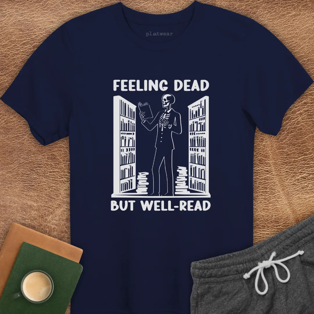 White crew neck t-shirt with the text 'FEELING DEAD BUT WELL-READ' printed on the front, displayed flat with a pair of gray shorts, a book, and a cup.