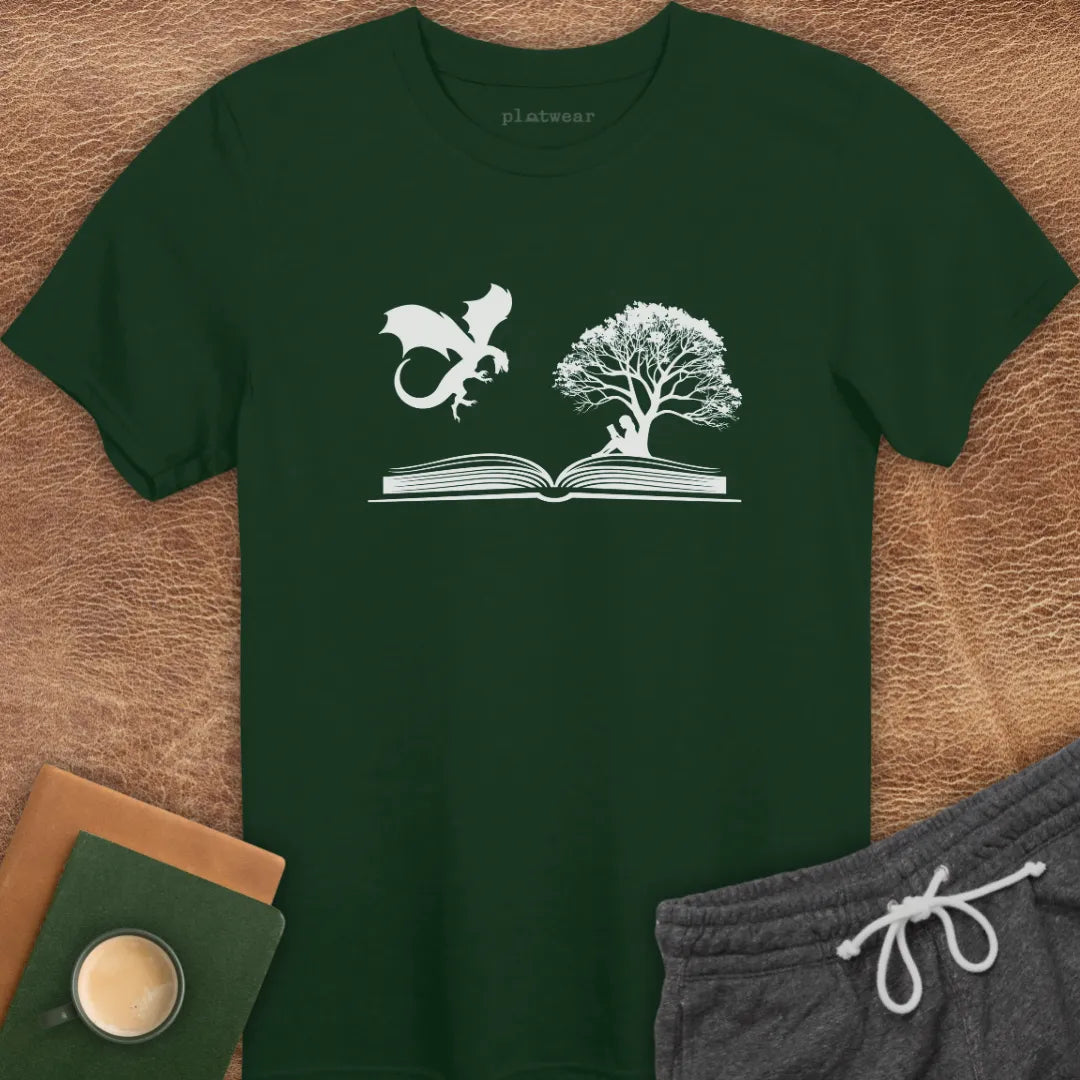 A gray T-shirt with a graphic print of a dragon and a tree on a book, positioned on a wooden surface with a notebook and a cup beside it.