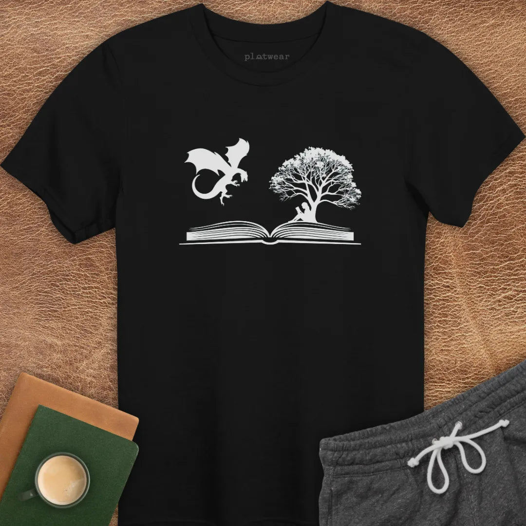 A gray T-shirt with a graphic print of a dragon and a tree on a book, positioned on a wooden surface with a notebook and a cup beside it.