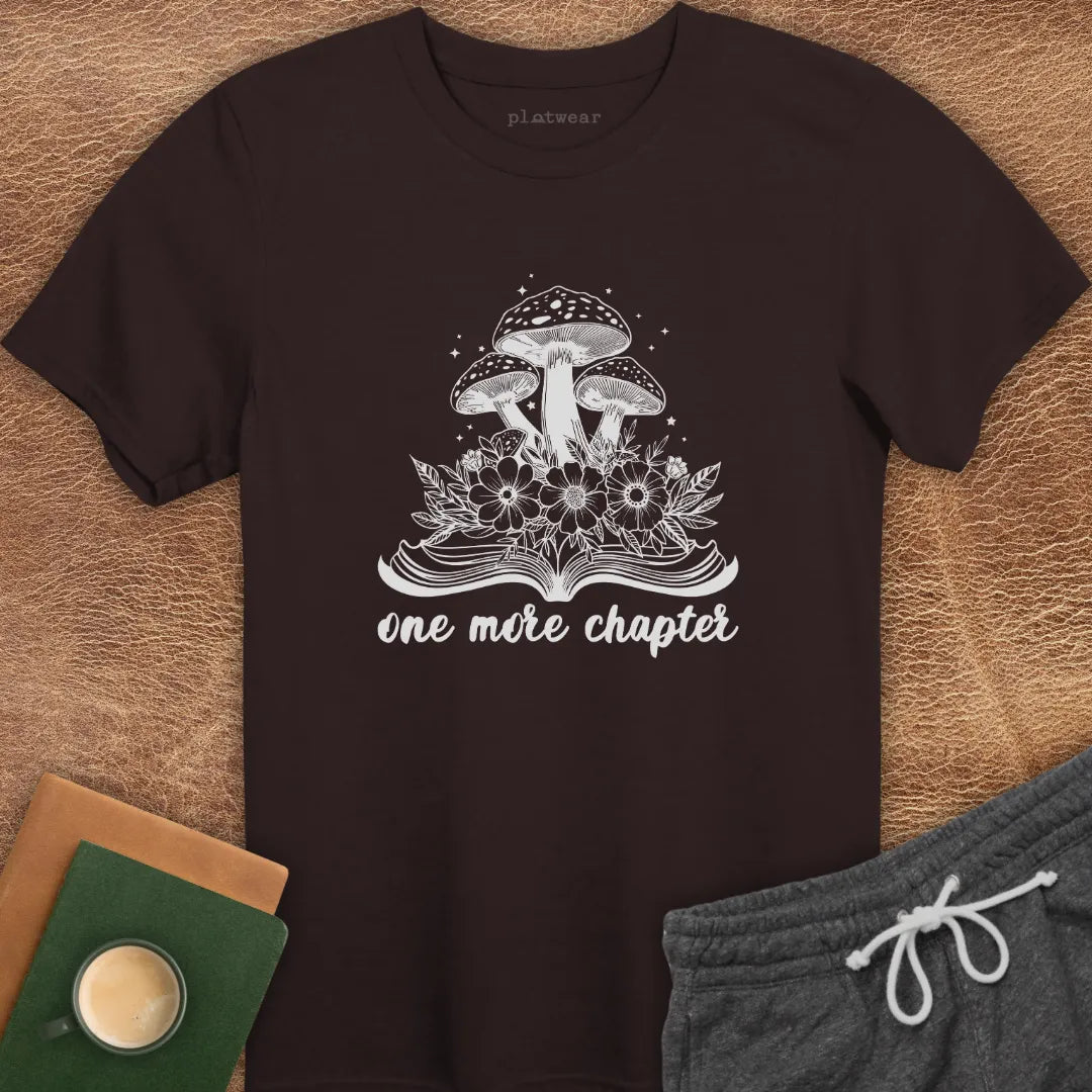 Maroon colored t-shirt with a graphic that includes a book and mushrooms, and the text 'one more chapter'. The shirt is presented flat with a pair of grey shorts and a cup on a wooden surface.