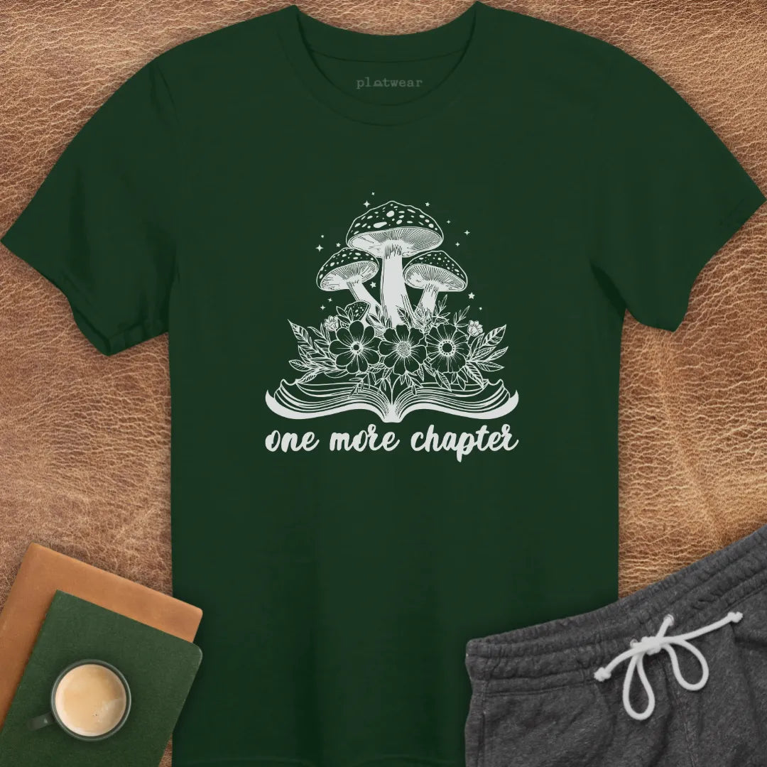 Maroon colored t-shirt with a graphic that includes a book and mushrooms, and the text 'one more chapter'. The shirt is presented flat with a pair of grey shorts and a cup on a wooden surface.