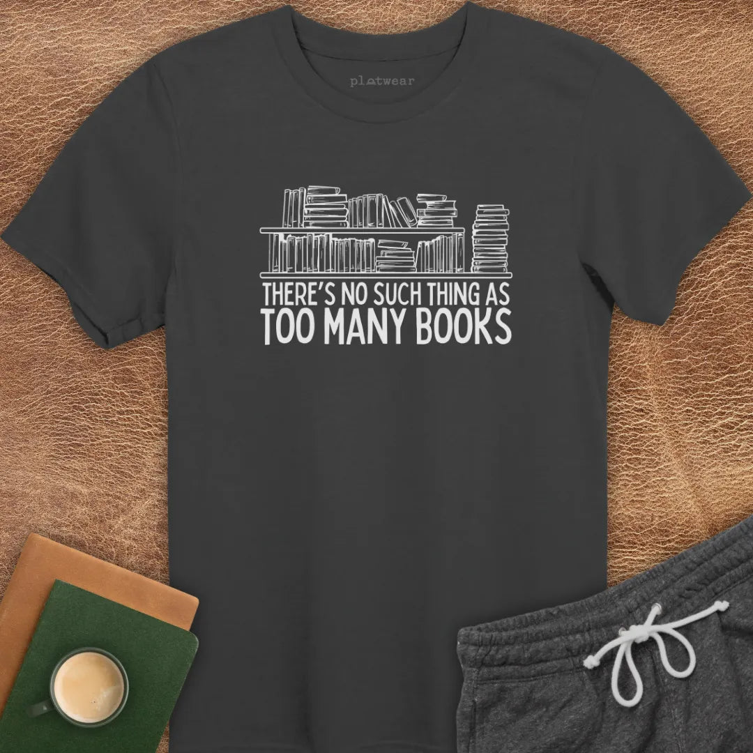 Black crew neck t-shirt with the text 'THERE'S NO SUCH THING AS TOO MANY BOOKS' printed on the front, displayed on a wooden surface alongside a book and a cup of coffee.