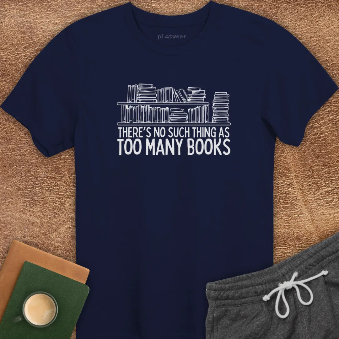 Black crew neck t-shirt with the text 'THERE'S NO SUCH THING AS TOO MANY BOOKS' printed on the front, displayed on a wooden surface alongside a book and a cup of coffee.