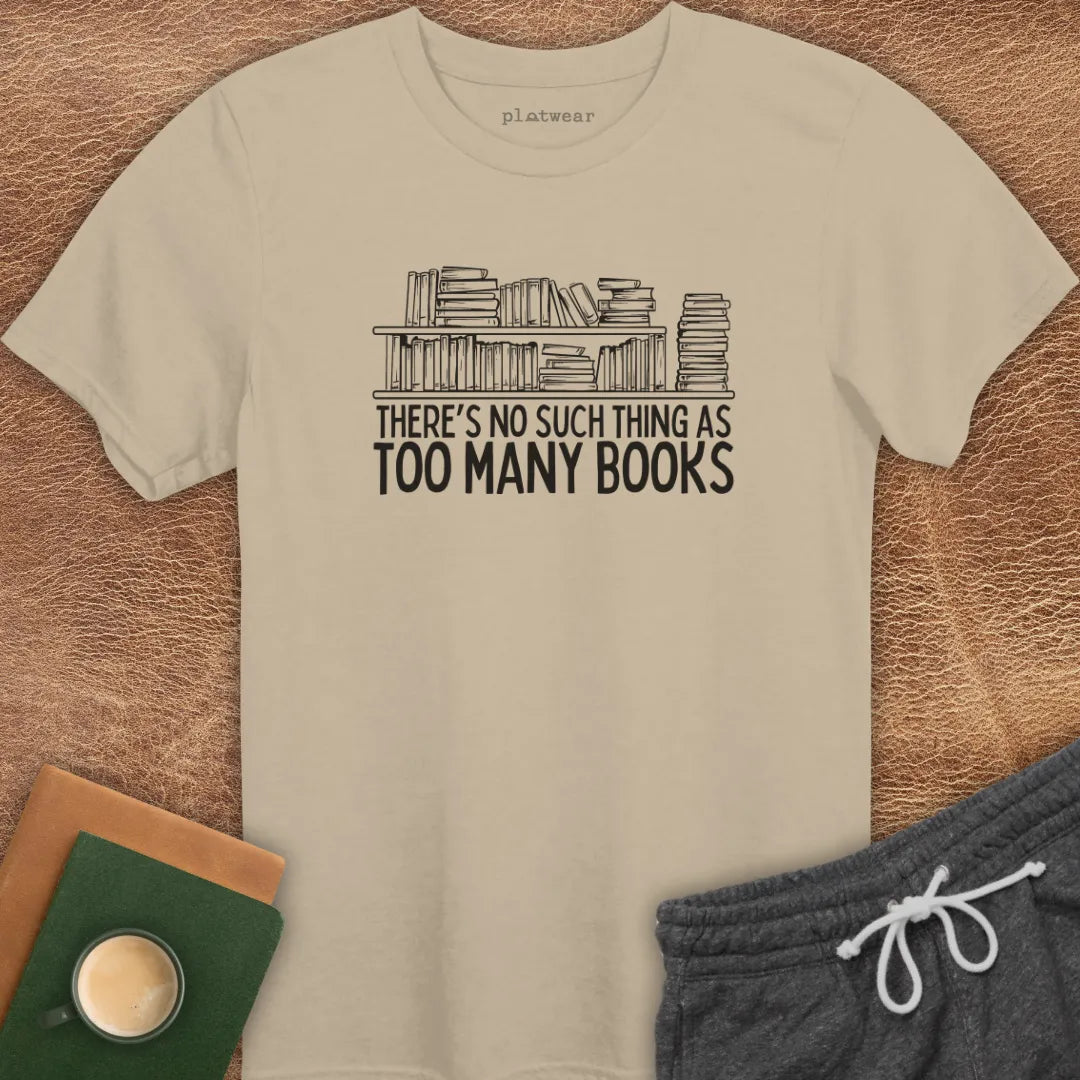 Black crew neck t-shirt with the text 'THERE'S NO SUCH THING AS TOO MANY BOOKS' printed on the front, displayed on a wooden surface alongside a book and a cup of coffee.