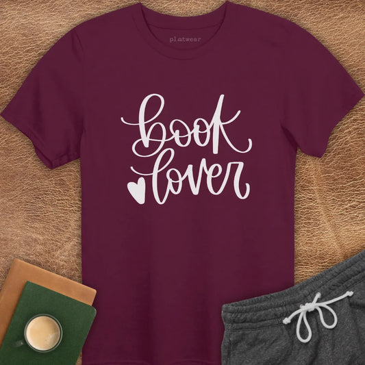 A maroon colored t-shirt with the text 'BOOK LOVER' and a heart symbol printed on the front, displayed flat with a pair of gray sweatpants and a book accessory items in the background.