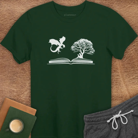 A gray T-shirt with a graphic print of a dragon and a tree on a book, positioned on a wooden surface with a notebook and a cup beside it.