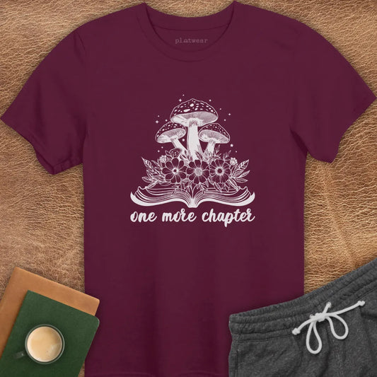 Maroon colored t-shirt with a graphic that includes a book and mushrooms, and the text 'one more chapter'. The shirt is presented flat with a pair of grey shorts and a cup on a wooden surface.