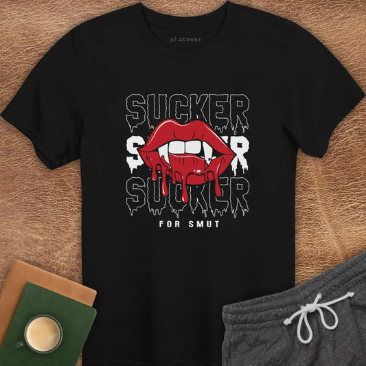 The image features a black crew neck t-shirt with the text "SUCKER FOR SMUT" printed in stylized red font, accompanied by a graphic of exaggerated red lips. The t-shirt is displayed flat on a brown surface, with a pair of gray sweatpants and a notebook in the background, suggesting a casual, everyday wear style.