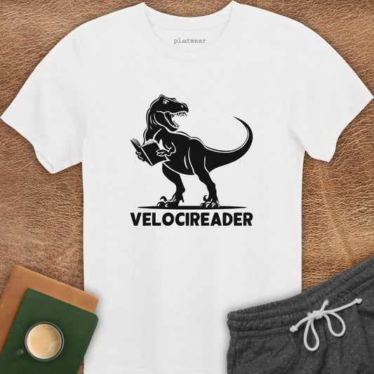 A white crew neck t-shirt with a graphic print of a dinosaur holding a book, and the word 'VELOCIREADER' printed below it. The t-shirt is displayed on a wooden surface with a book, a notebook, and a cup nearby.